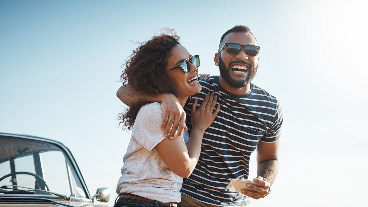 A couple laughing while standing next to a car on the beach.