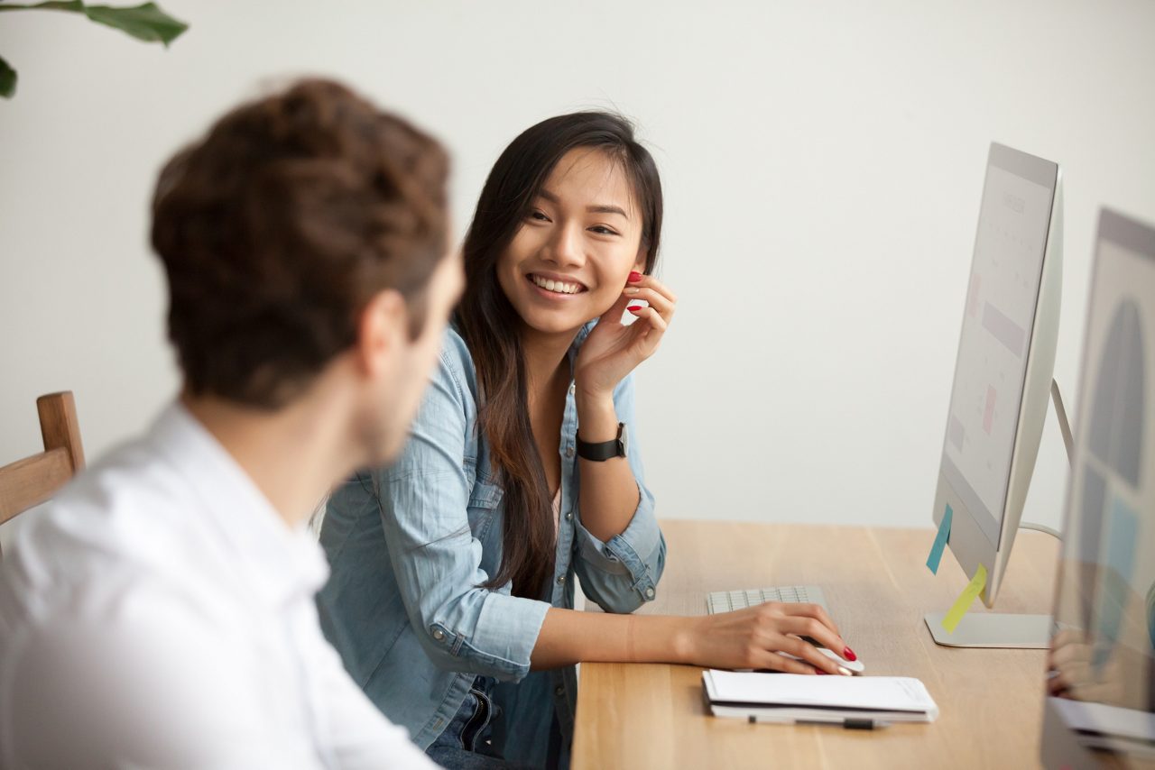 Smiling woman in front of computer talking to male colleague at work sharing office desk with desktops