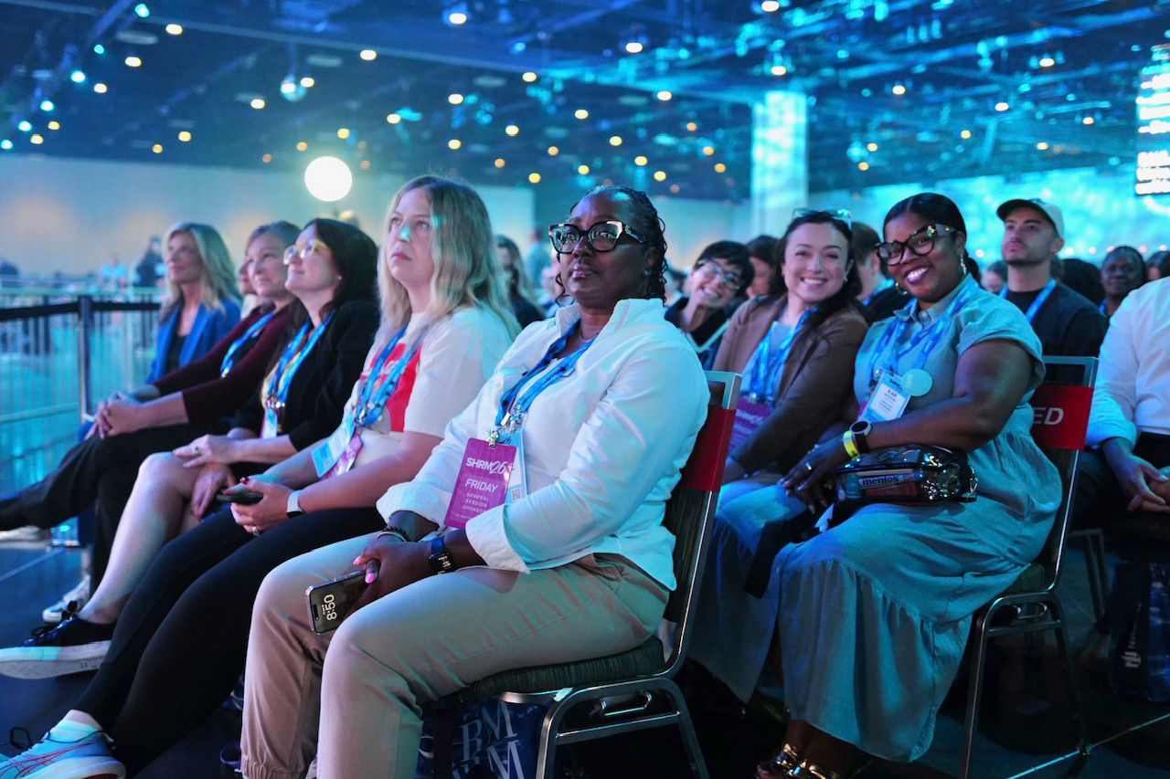 A crowd of smiling SHRM26 attendees in business casual attire look attentively toward the stage. 