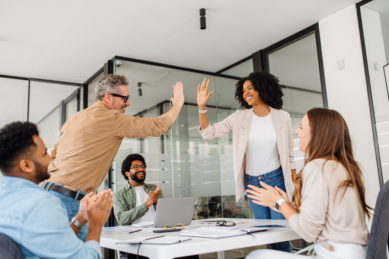 A diverse team of professionals celebrates a workplace achievement with a high-five in a modern office. The moment captures motivation, cooperation, and a positive business culture focused on teamwork