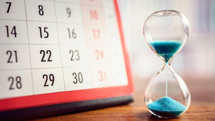 An hourglass with blue sand in front of a calendar.