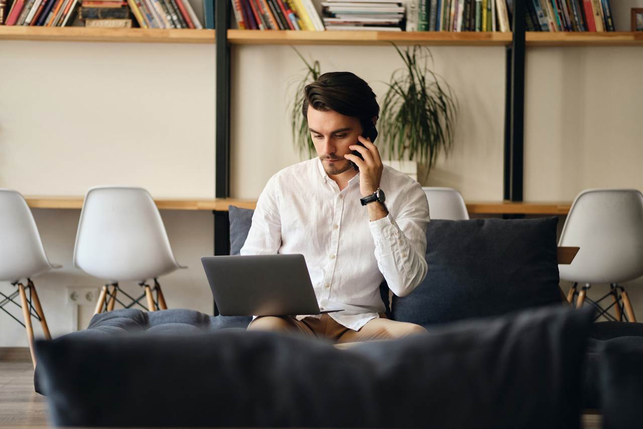 Young handsome businessman sitting on sofa thoughtfully talking on cellphone and working on laptop in modern co-working space