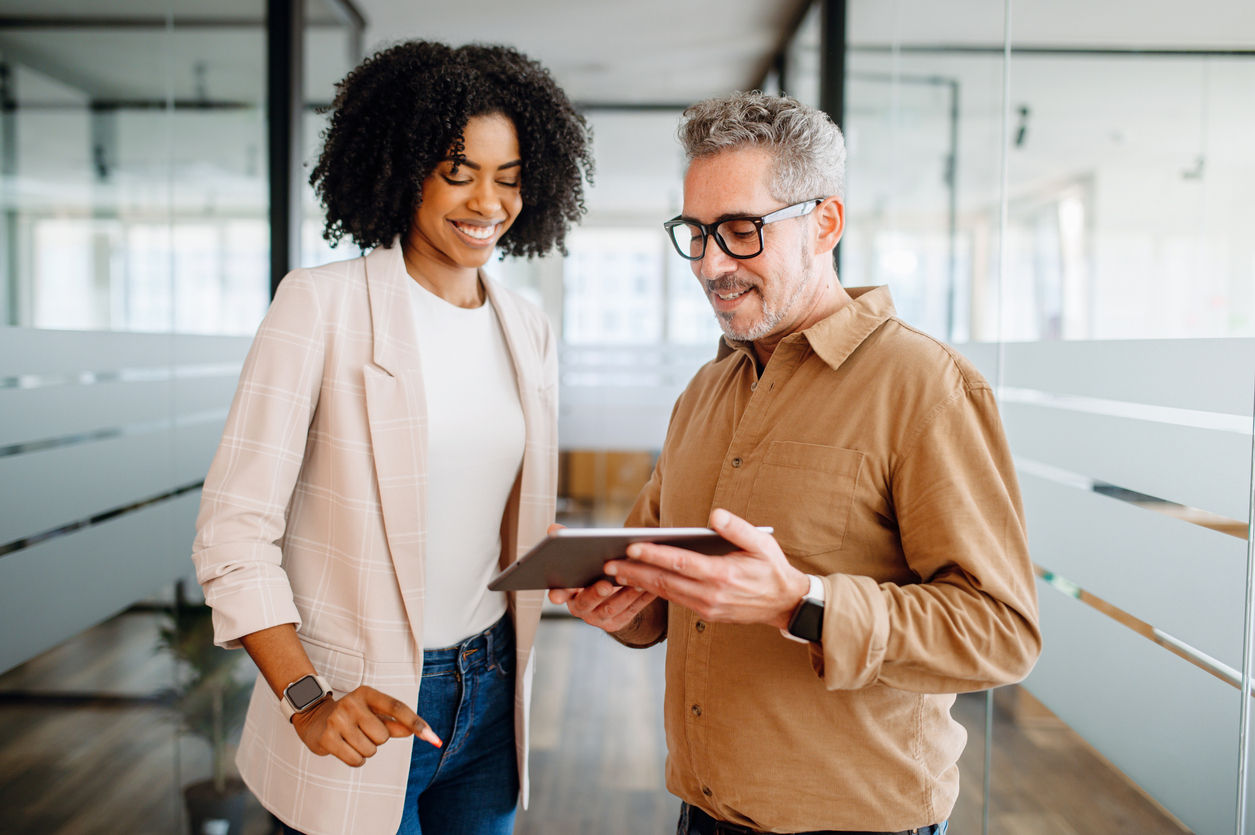 A businesswoman and a male colleague collaboratively review content on a tablet in a bright office space, reflecting a multigenerational workplace