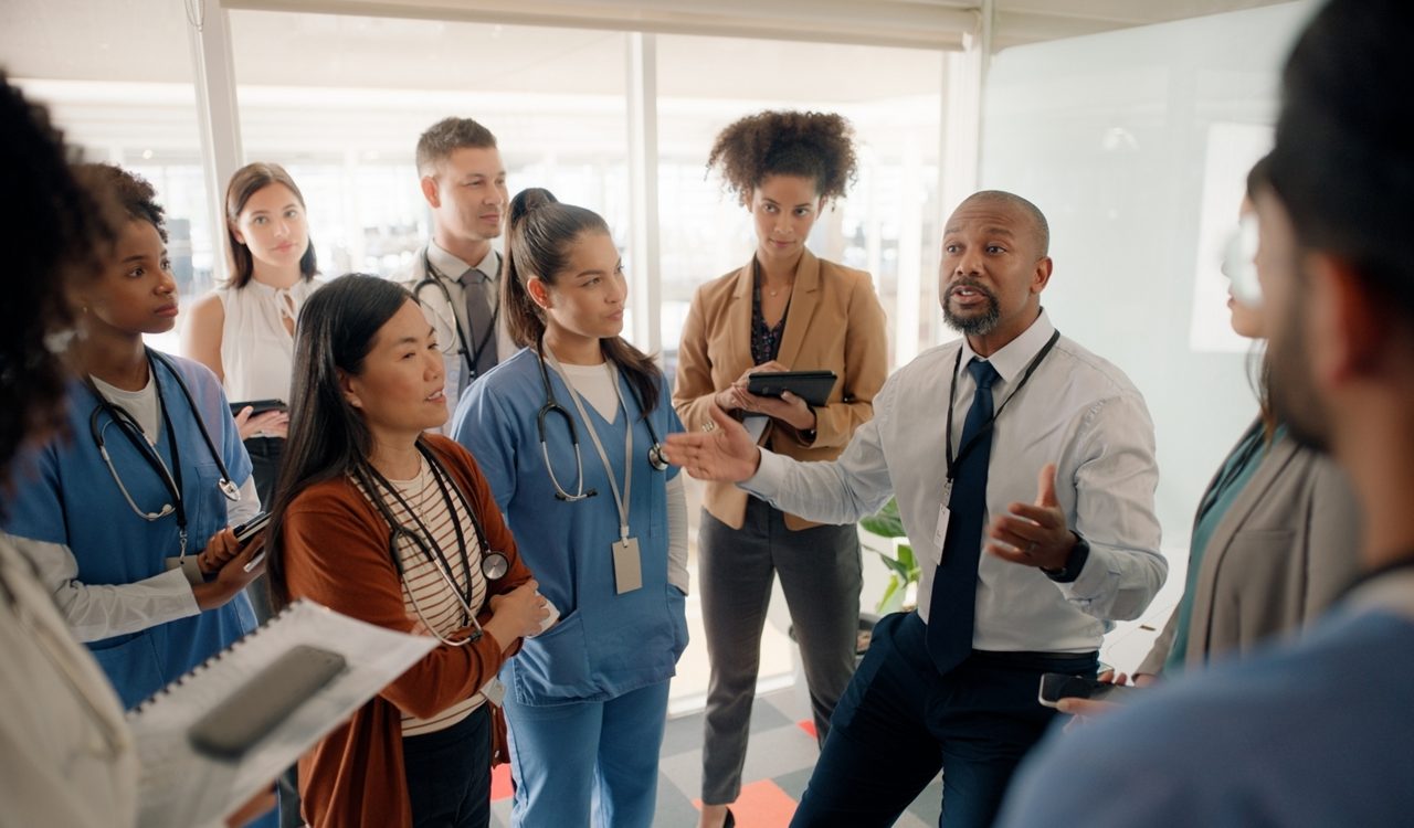 Boardroom, discussion and motivation for staff by executive in hospital for healthcare. Man, training and mentor of employees or nurses and tablets for notes of speech by leader in medical field