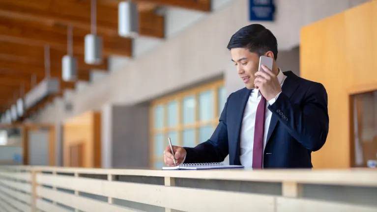 A man in a blue suit and red tie talks on a smartphone while overlooking an internal atrium railing on the top floor of an office building.