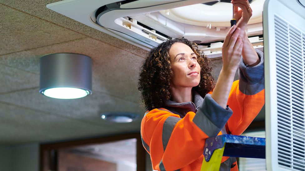 A woman on a ladder working on an air conditioning unit.