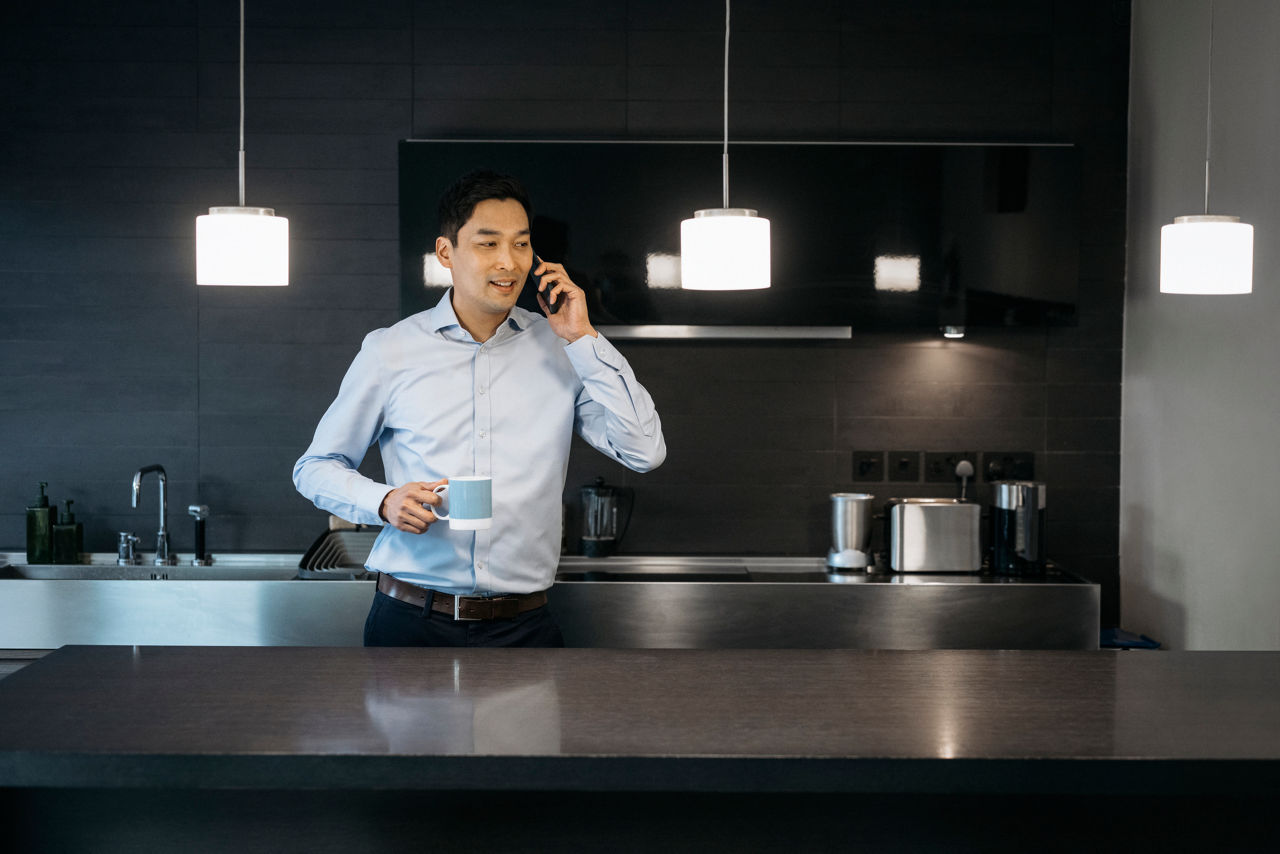 Waist-up view of smiling man in relaxed business attire standing in modern domestic kitchen with cup of coffee and talking on smart phone.