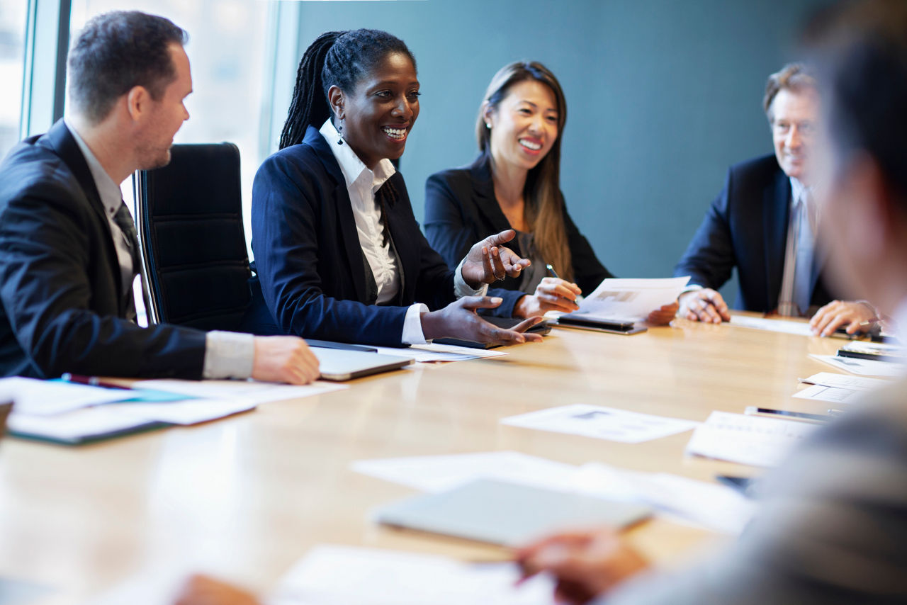A black businesswoman discusses plans during a meeting with a multiracial group of work colleagues in businesswear at a board room conference table.