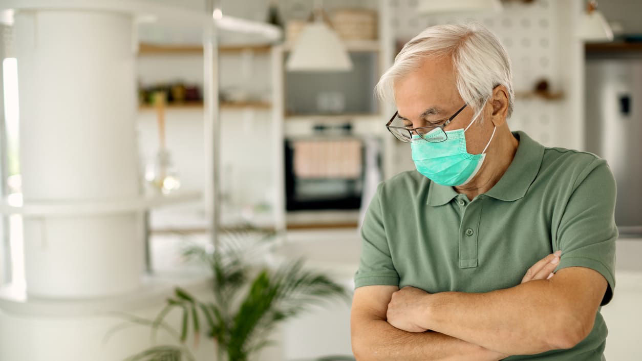 An elderly man wearing a surgical mask in his home.