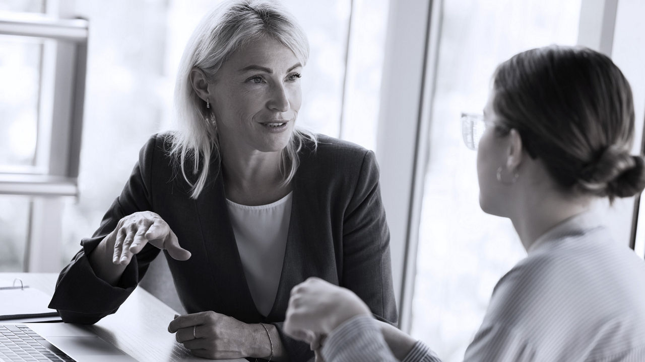 A blonde woman in a blazer and white top provides constructive feedback to a woman with brown hair and glasses, who sits with her back to the camera.