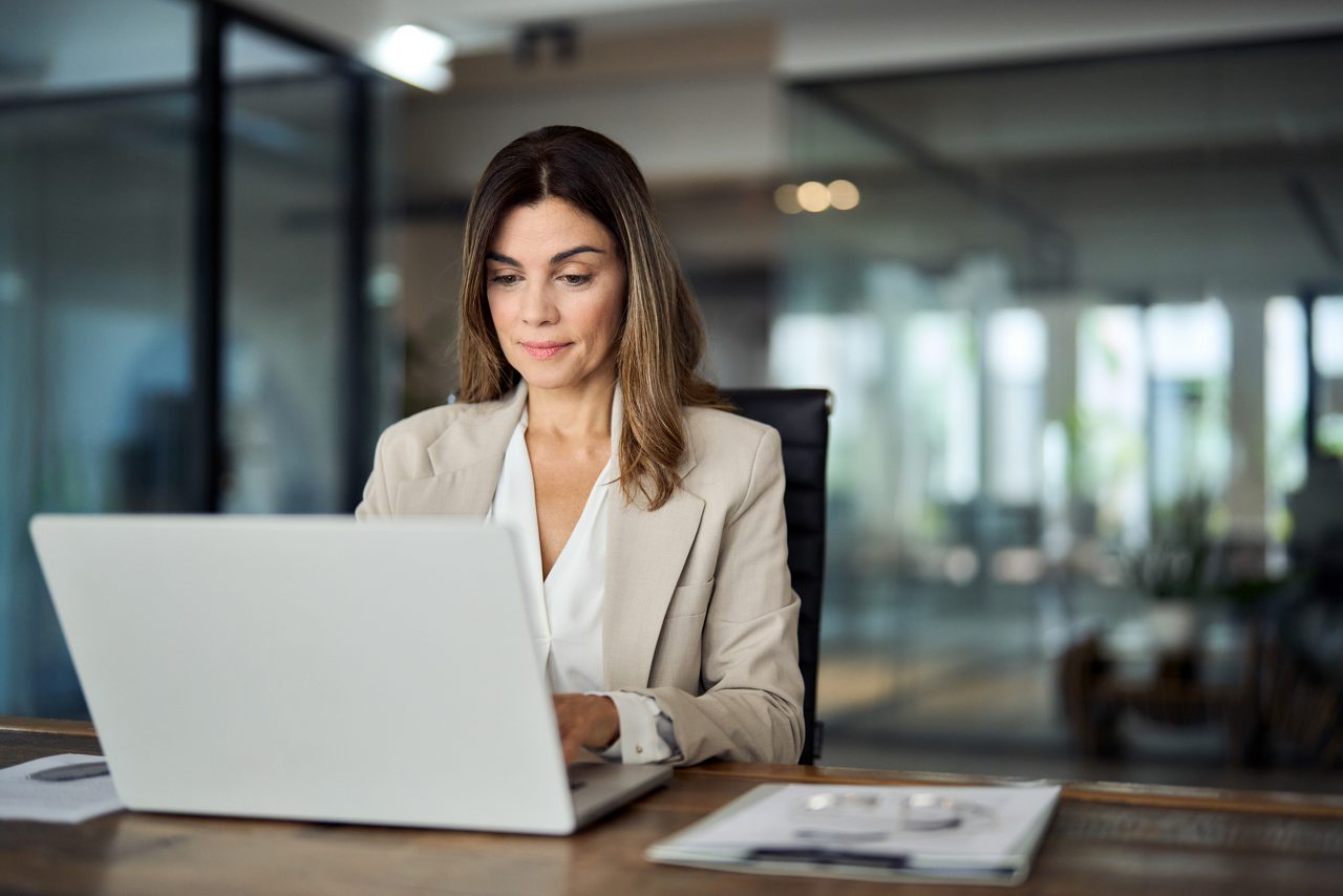 Busy mature middle aged professional business woman manager executive looking at laptop computer technology in office working on digital financial banking market sitting at desk. Copy space.