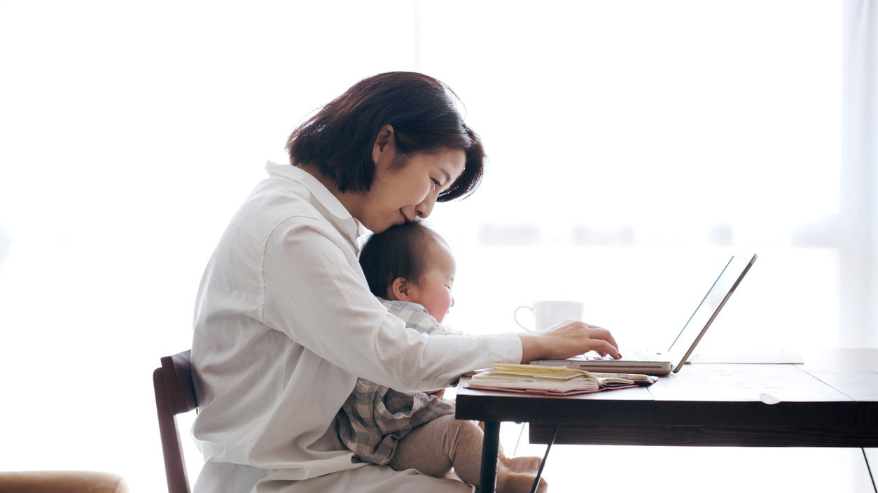 A woman is using a laptop while holding a baby.