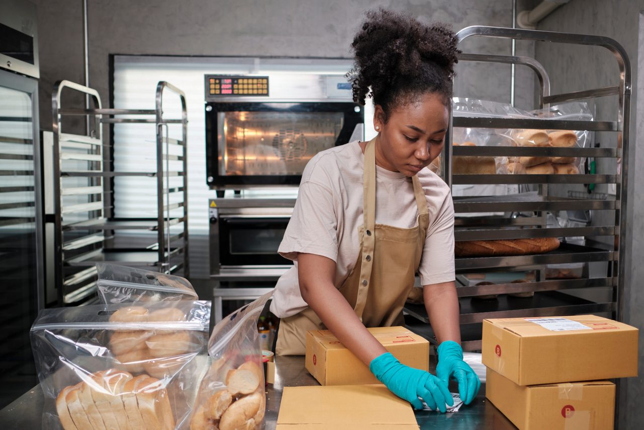 Bakery startup small business delivery. One African American female cook is packing handmade and fresh-baked bread and pastries in boxes and sending for online customer purchases in culinary kitchen.