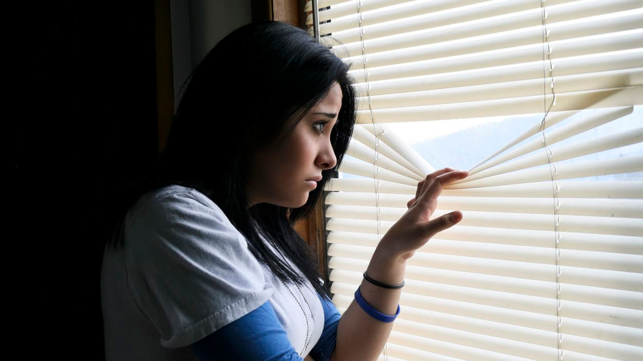 A woman looking out of a window with blinds.