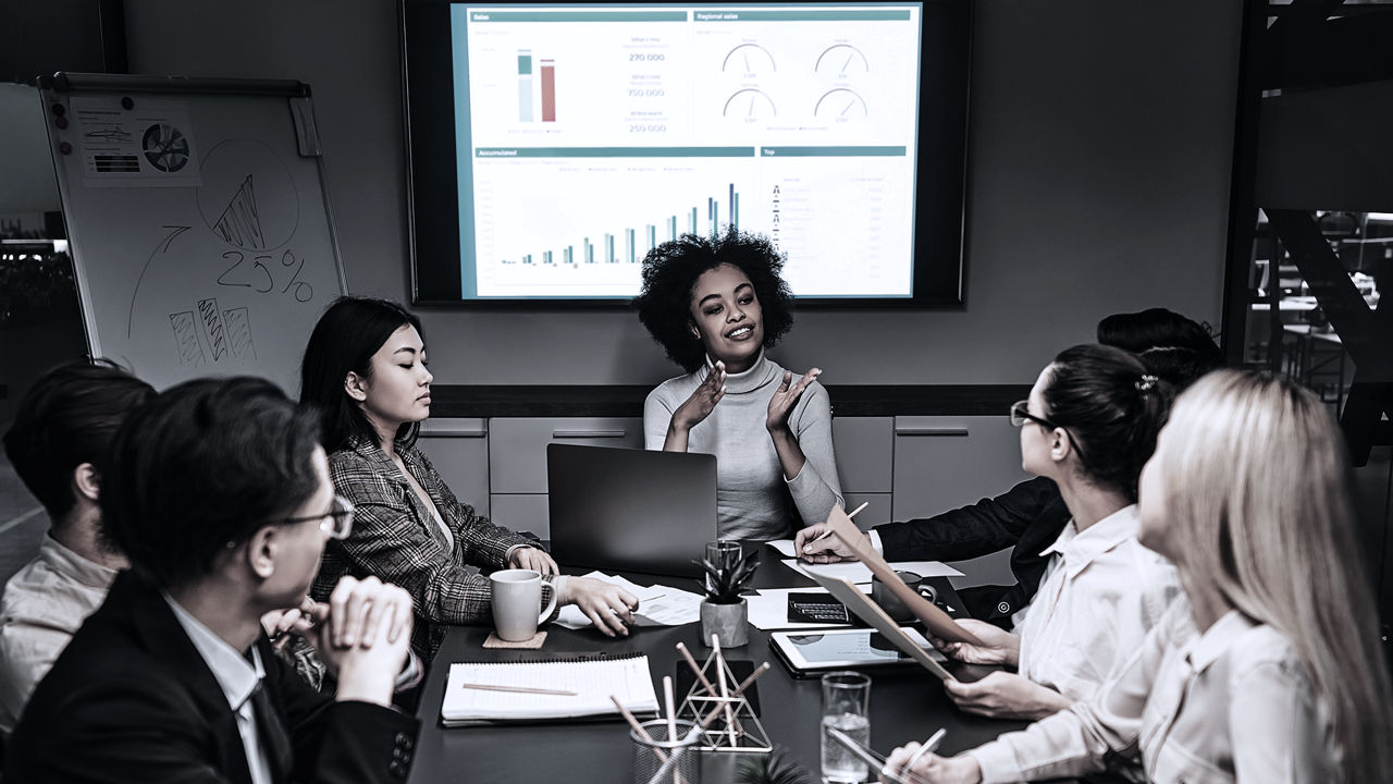 Female leader presenting to team, all sitting around table with data on screen behind them