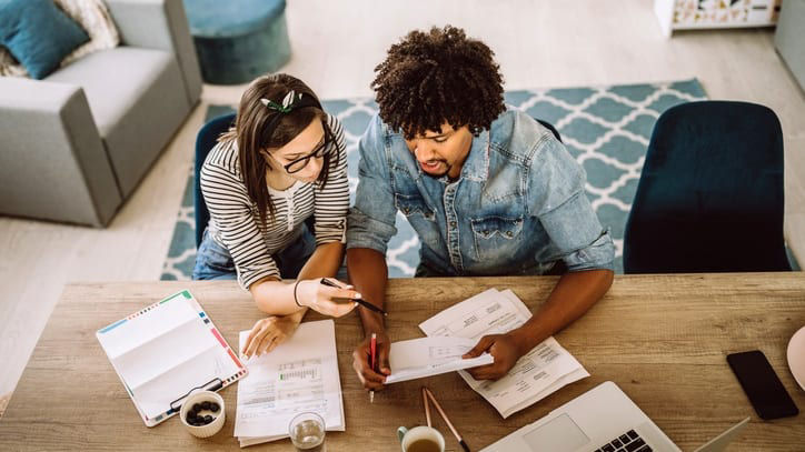 Two coworkers review financial documents together at a table with papers, a laptop, and notebooks, collaborating on budgeting or planning tasks.