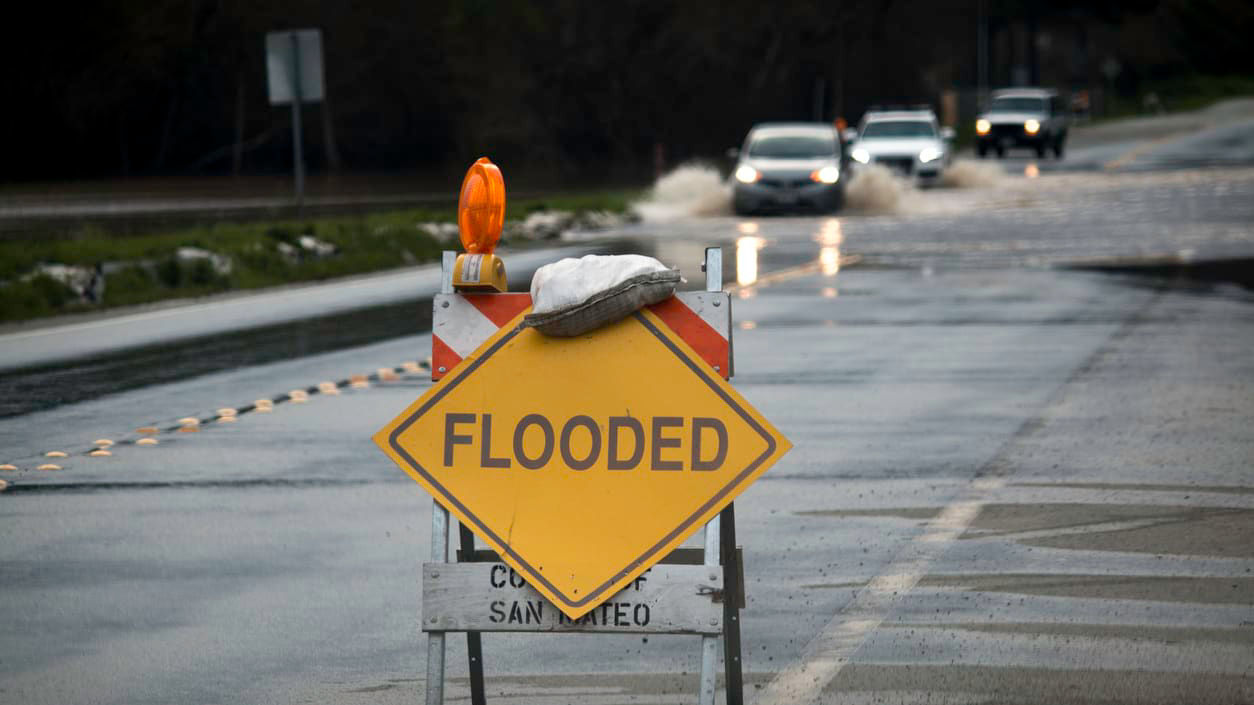 A flood warning sign sits on the side of a flooded road.