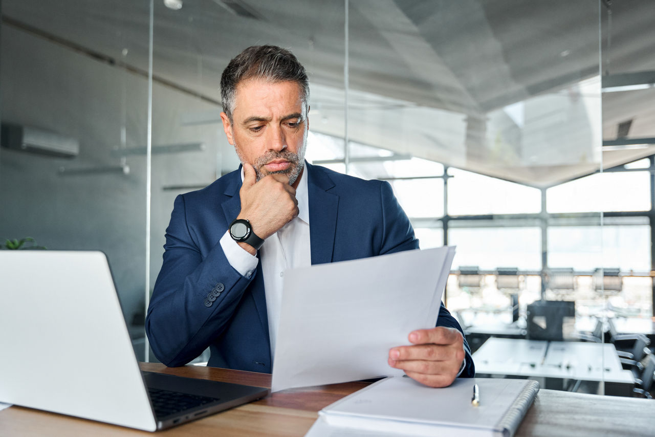 mid-age male office worker sitting behind desk with laptop open holding piece of paper