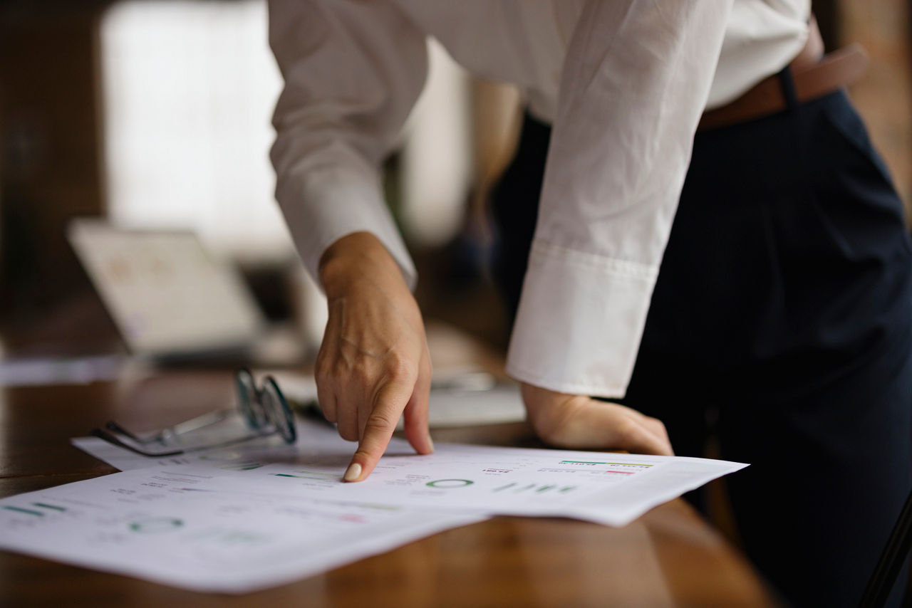 A woman in smart casual attire reviews printed charts on a desk with glasses nearby, indicating focused work in a modern office environment. A blurred laptop is visible in the background.