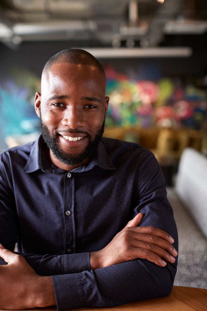 Mid adult black male creative sitting at a table in an office cafe smiling to camera, close up, crop