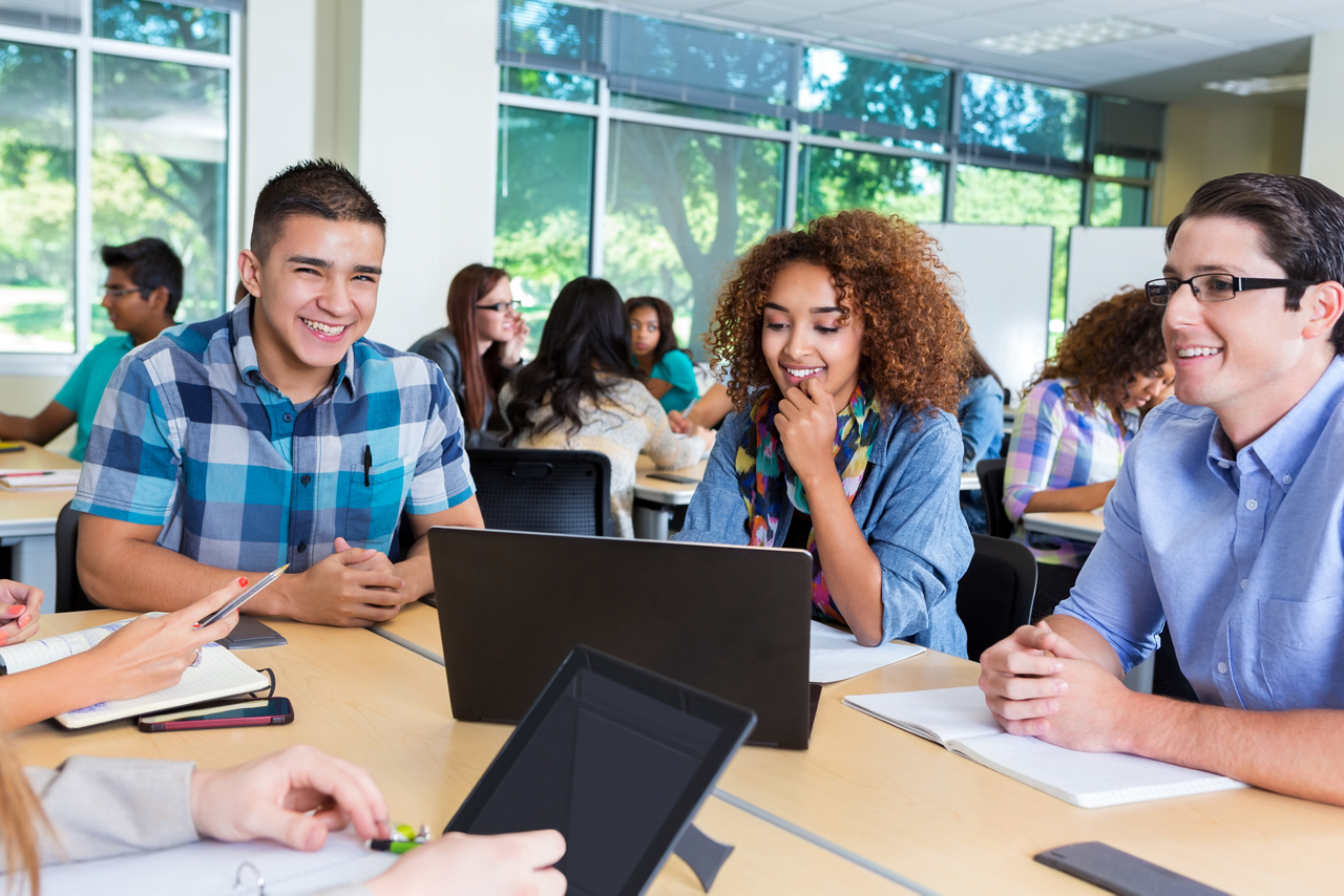 Hispanic, African American, and Caucasian teens or young adults are studying together while sitting in high school or college classroom. They are using laptop computer to study for class assignment. Students are working at desks in background.