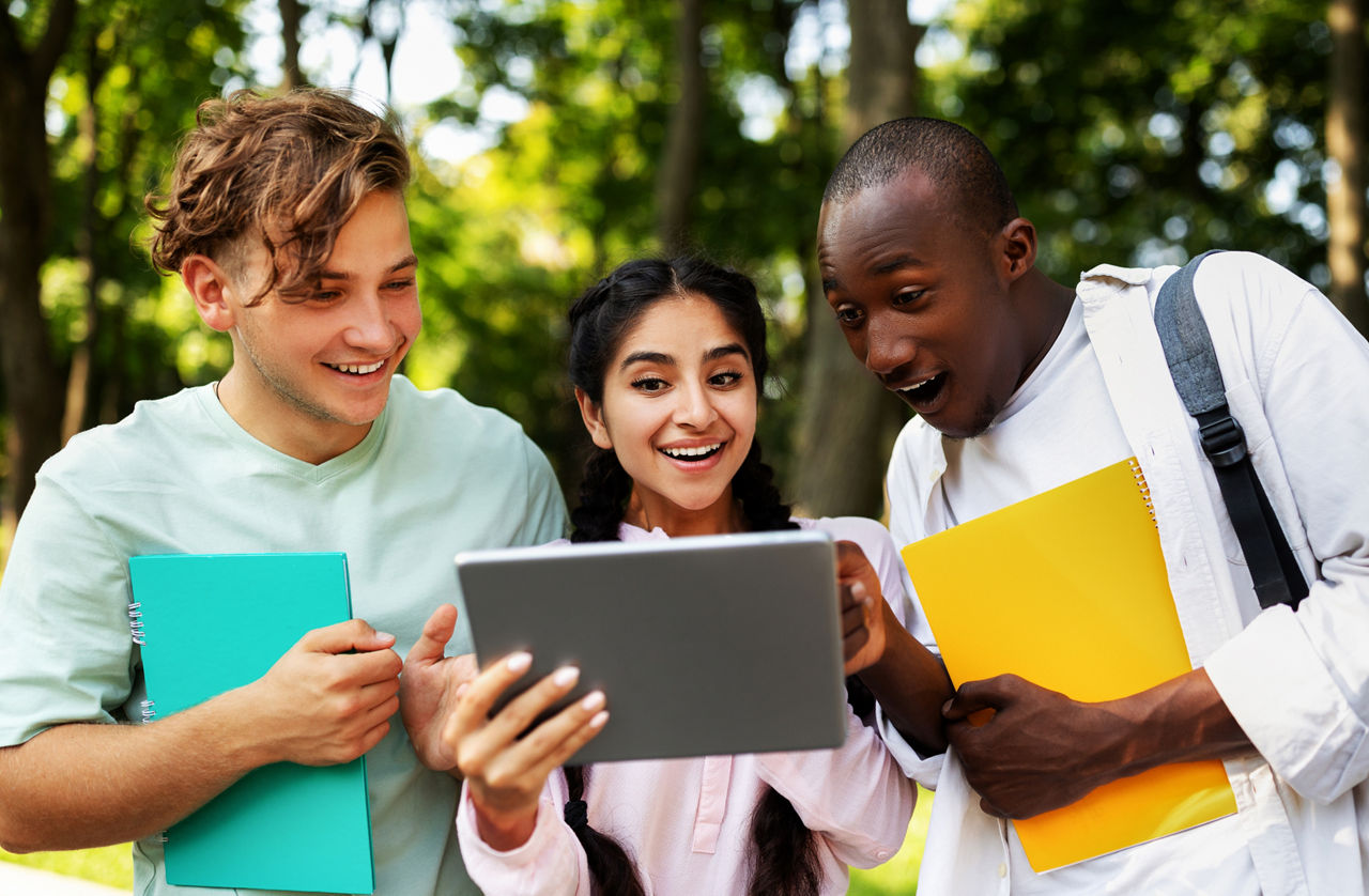 Three teens outdoors smiling and reacting to a tablet, holding notebooks and backpacks, appearing excited and engaged in learning together.