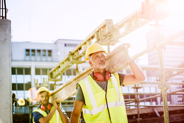 Male construction workers in protective workwear carrying plank at site