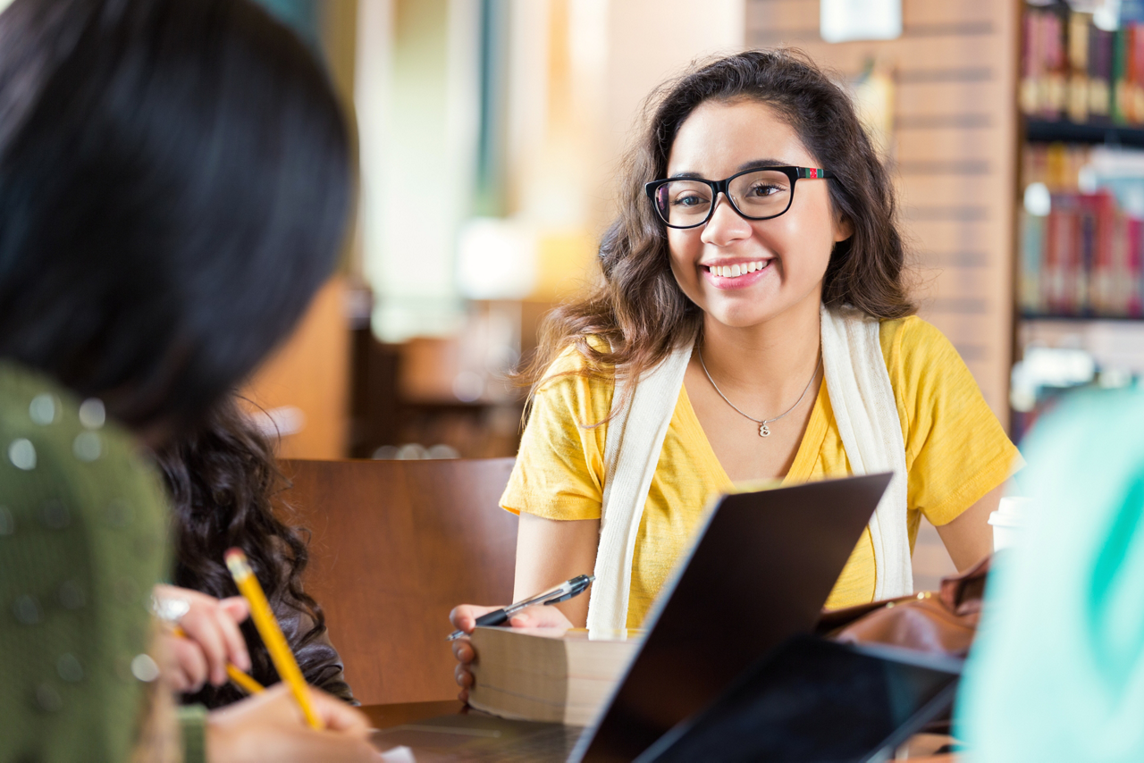 Trendy hipster Hispanic college age girl is smiling while sitting at tabe in library with group of friends. Students are working in study group to prepare for exam while using laptop computers, digital tablets, and other technology.