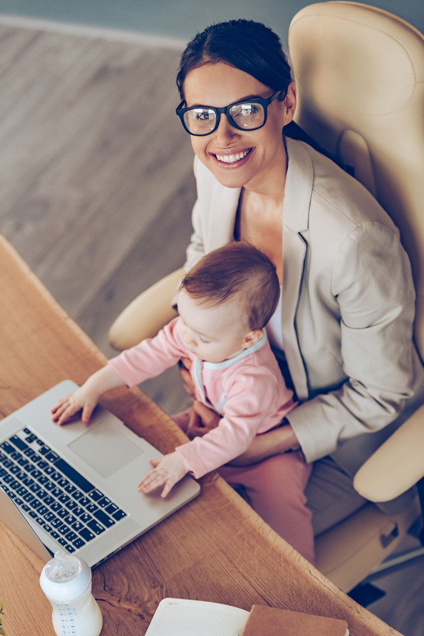 Top view of young beautiful businesswoman looking at camera with smile while sitting with baby girl on her knees at her working place