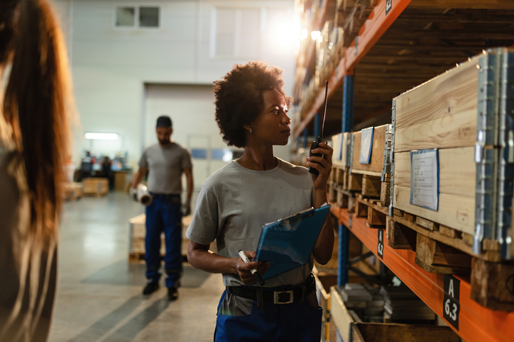Black female dispatcher checking stock of products in delivery warehouse and communicating on walkie-talkie.