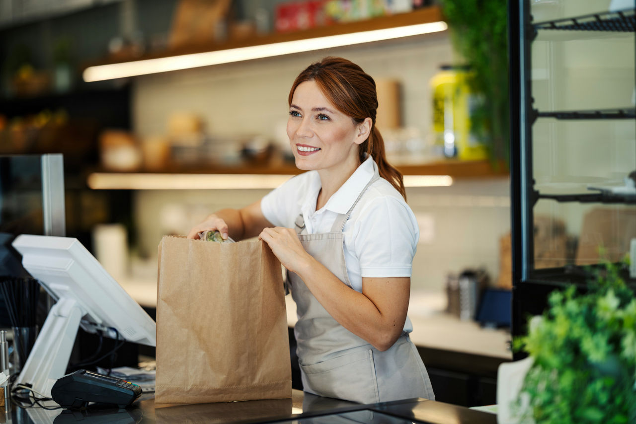 Woman cashier smiling while packing items into a brown paper shopping bag at a modern grocery store checkout counter