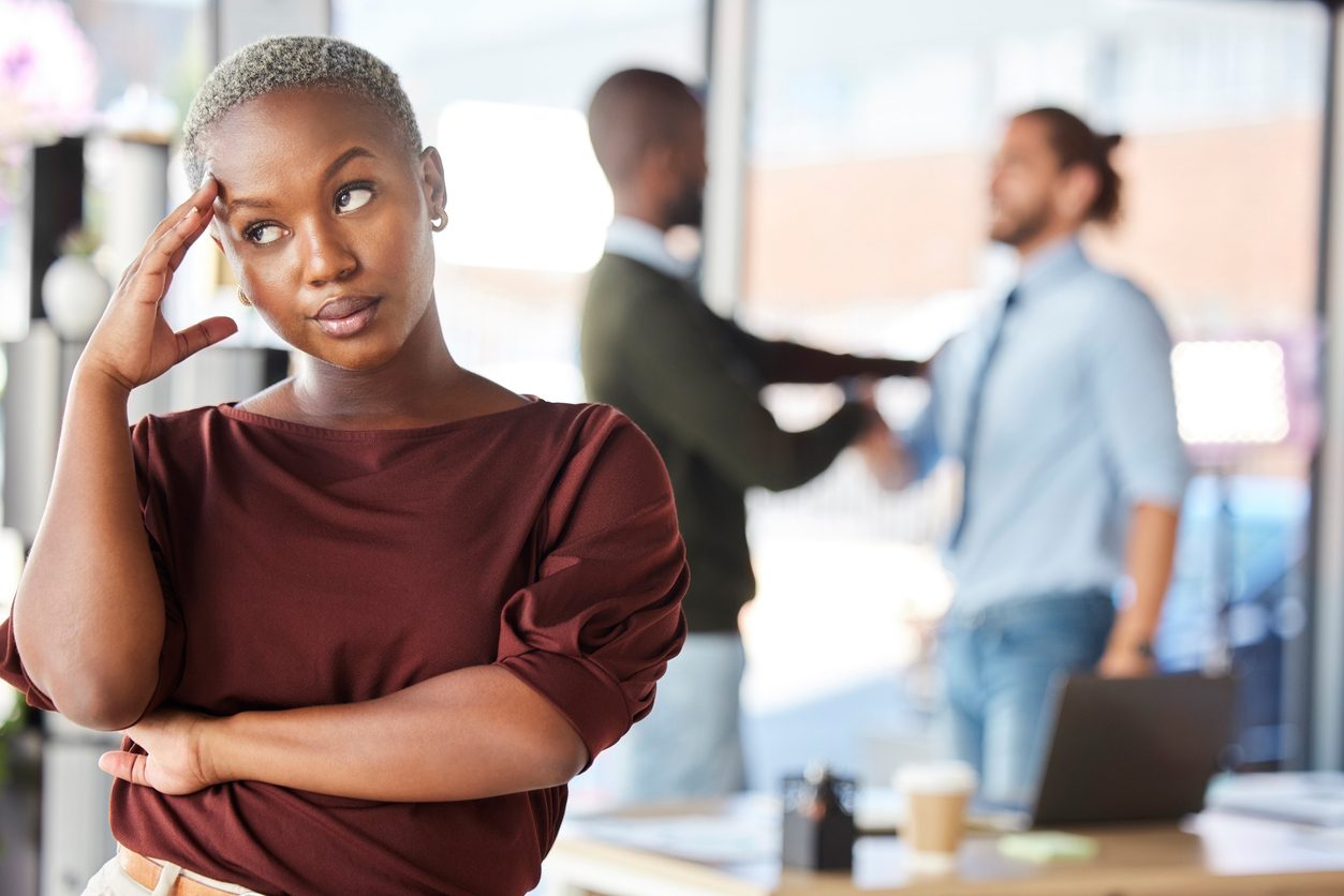 woman looking annoyed in an office environment, with two men shaking hands behind her.