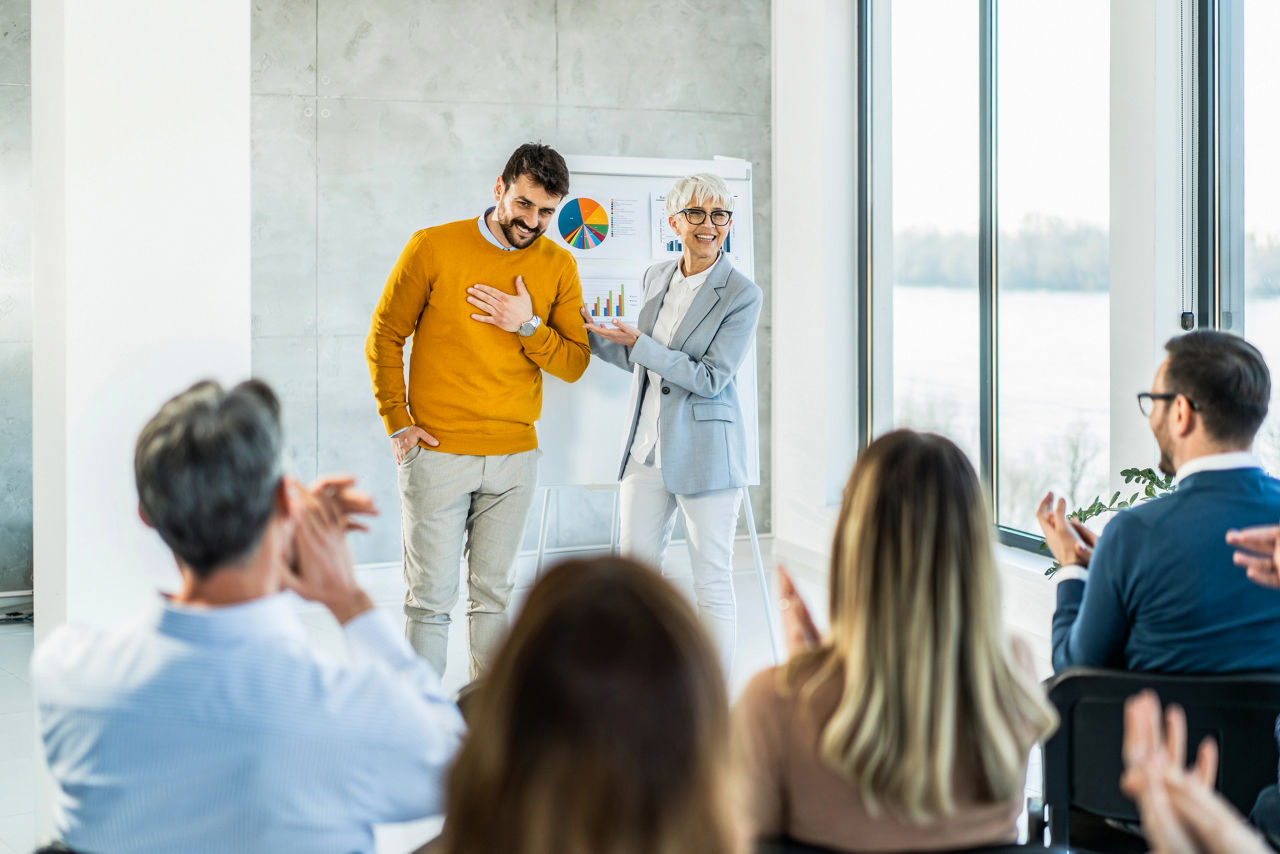 Happy senior businesswoman introducing a new young member of a company to a group of business people. Business people congratulating a applauding to young man for job well done! Young man being introduced to his new colleagues.