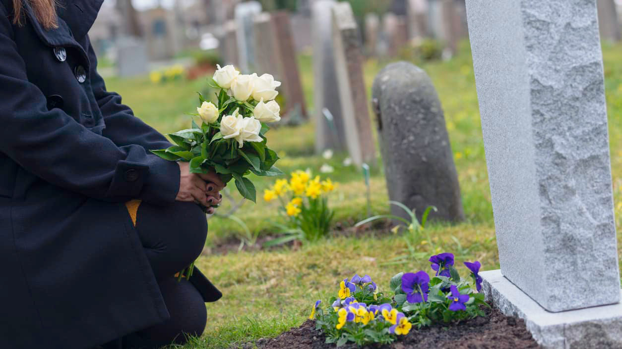 A woman kneeling down next to a grave with flowers.