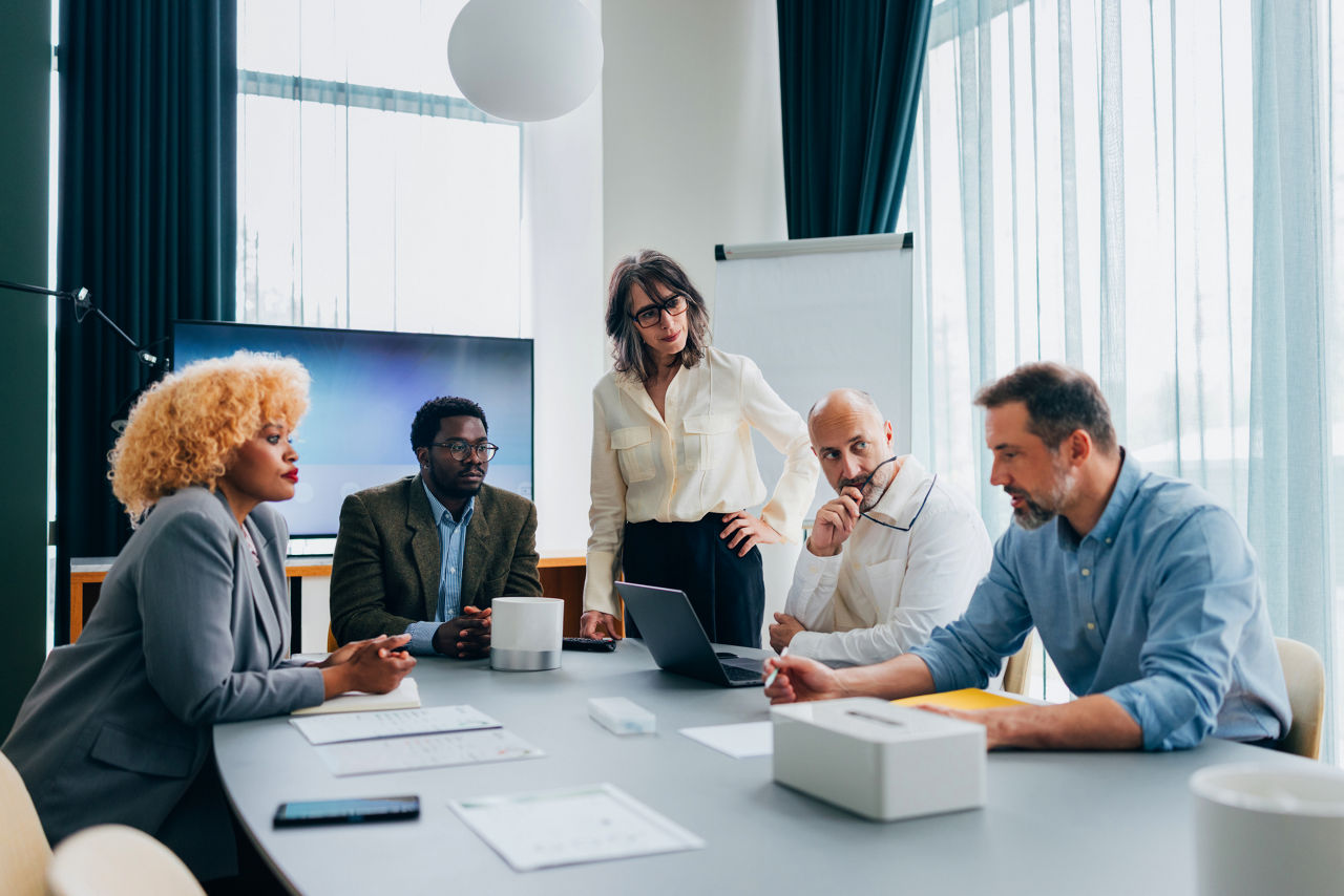 A group of professionals collaborates in a business meeting, brainstorming ideas and solutions in a corporate office environment. The diverse team appears focused and engaged in a strategic discussion around a table.