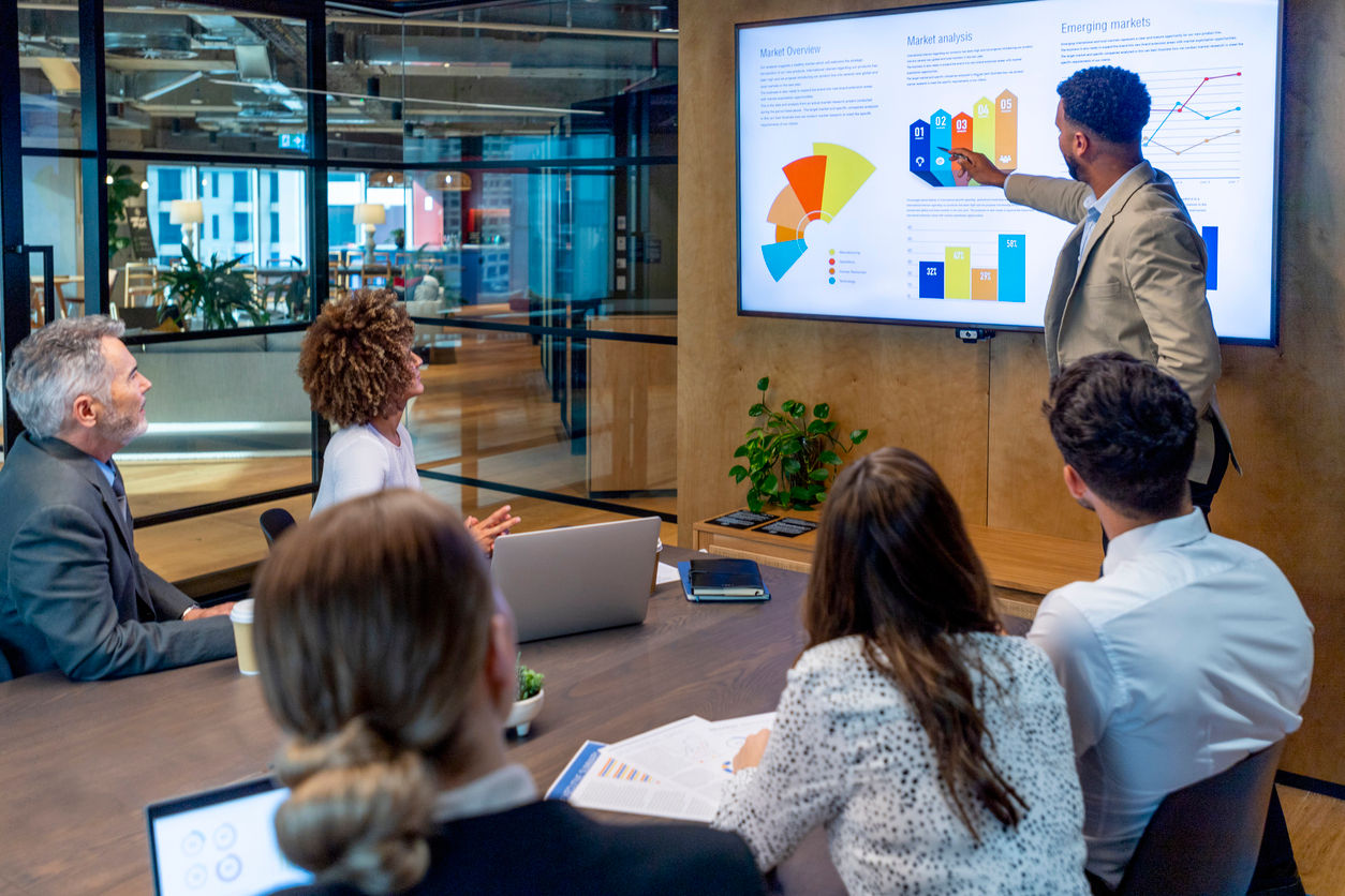 A man gives a big data presentation on a big screen in a board room. There are several financial graphs and charts on the screen with a diverse group of people in the meeting room. There is paperwork and technology on the table.