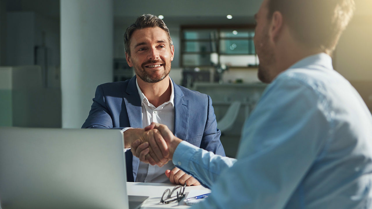 Two businessmen shaking hands in front of a laptop.