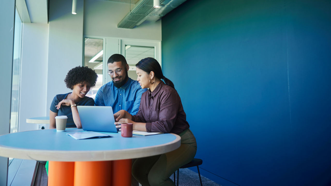 Three professionals working together seated around a small round table before an open laptop.