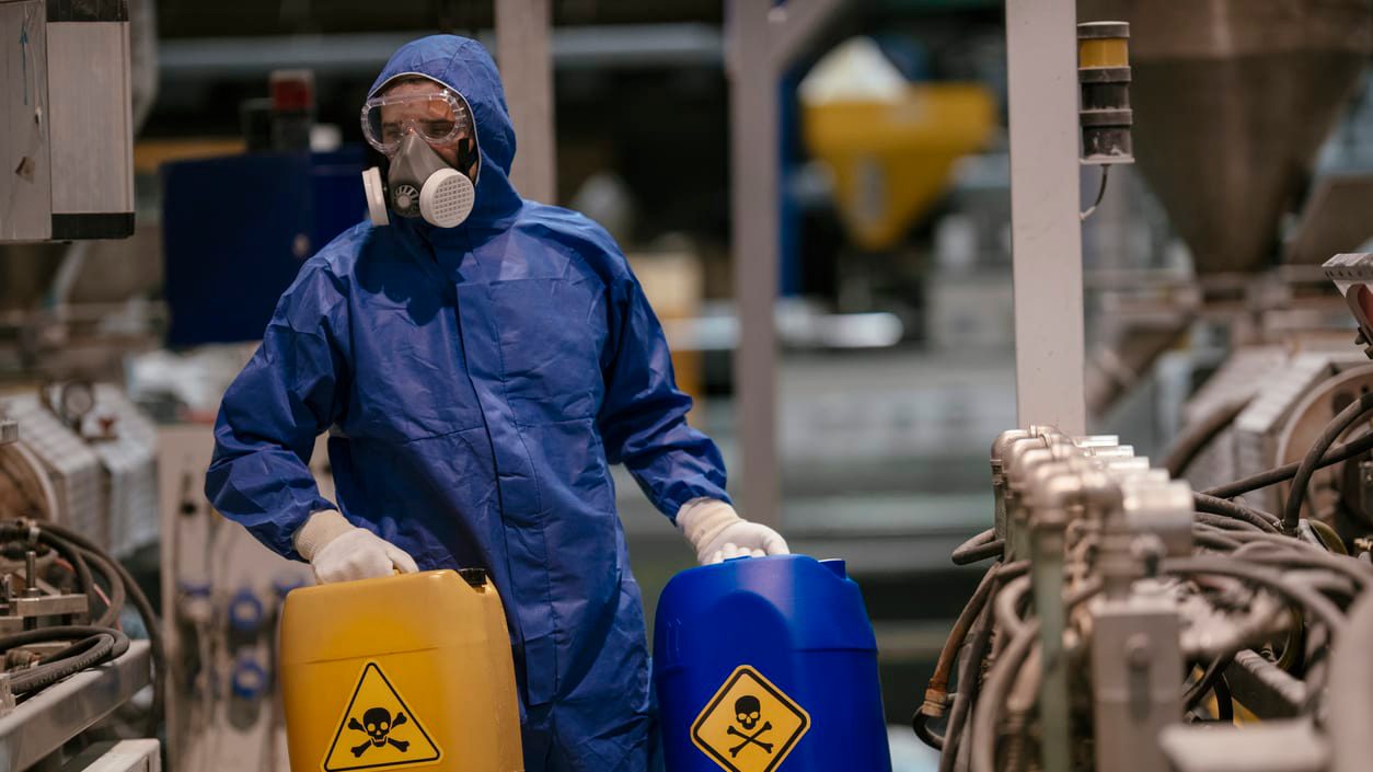 A worker in a protective suit is holding a bucket of chemicals.