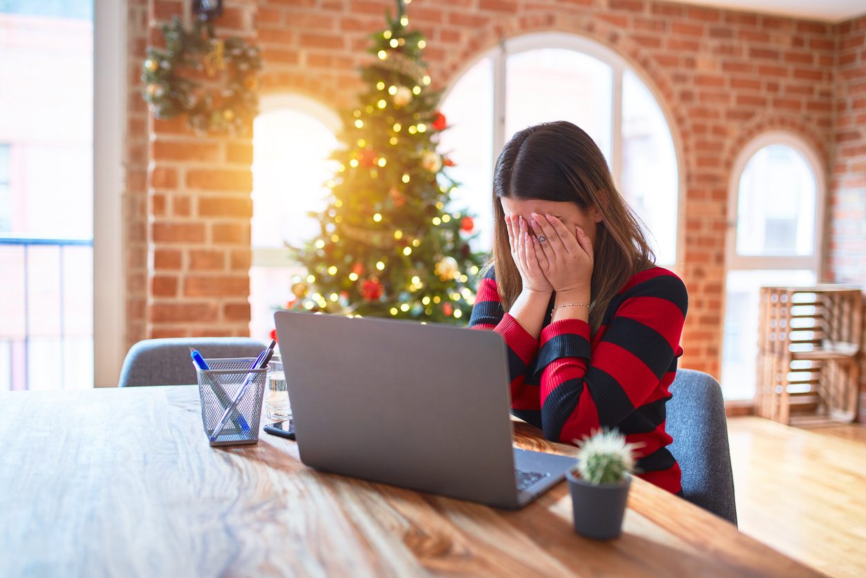 woman by a Christmas tree working on laptop with her face in her hands, indicating stress