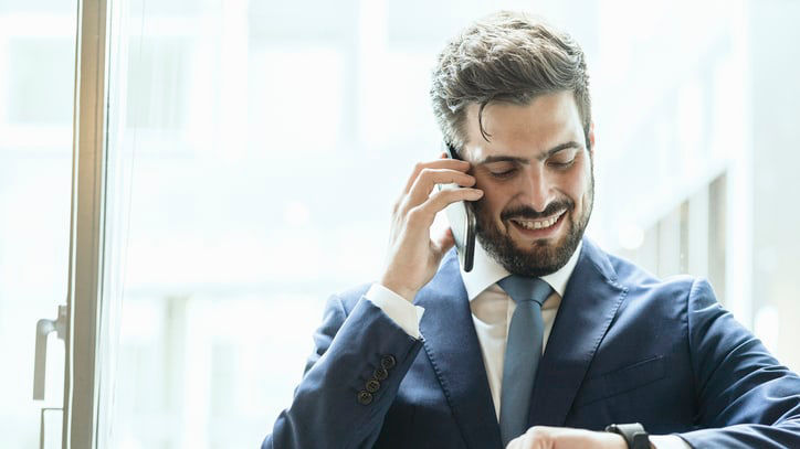 A man in a suit is talking on the phone while looking at his watch.