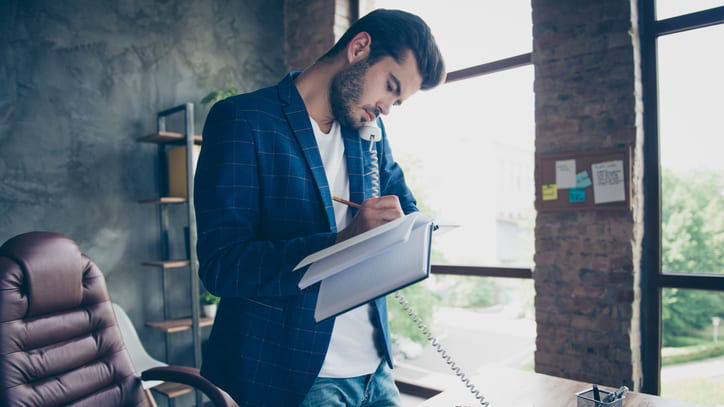 A man is writing in a notebook while sitting at a desk in an office.