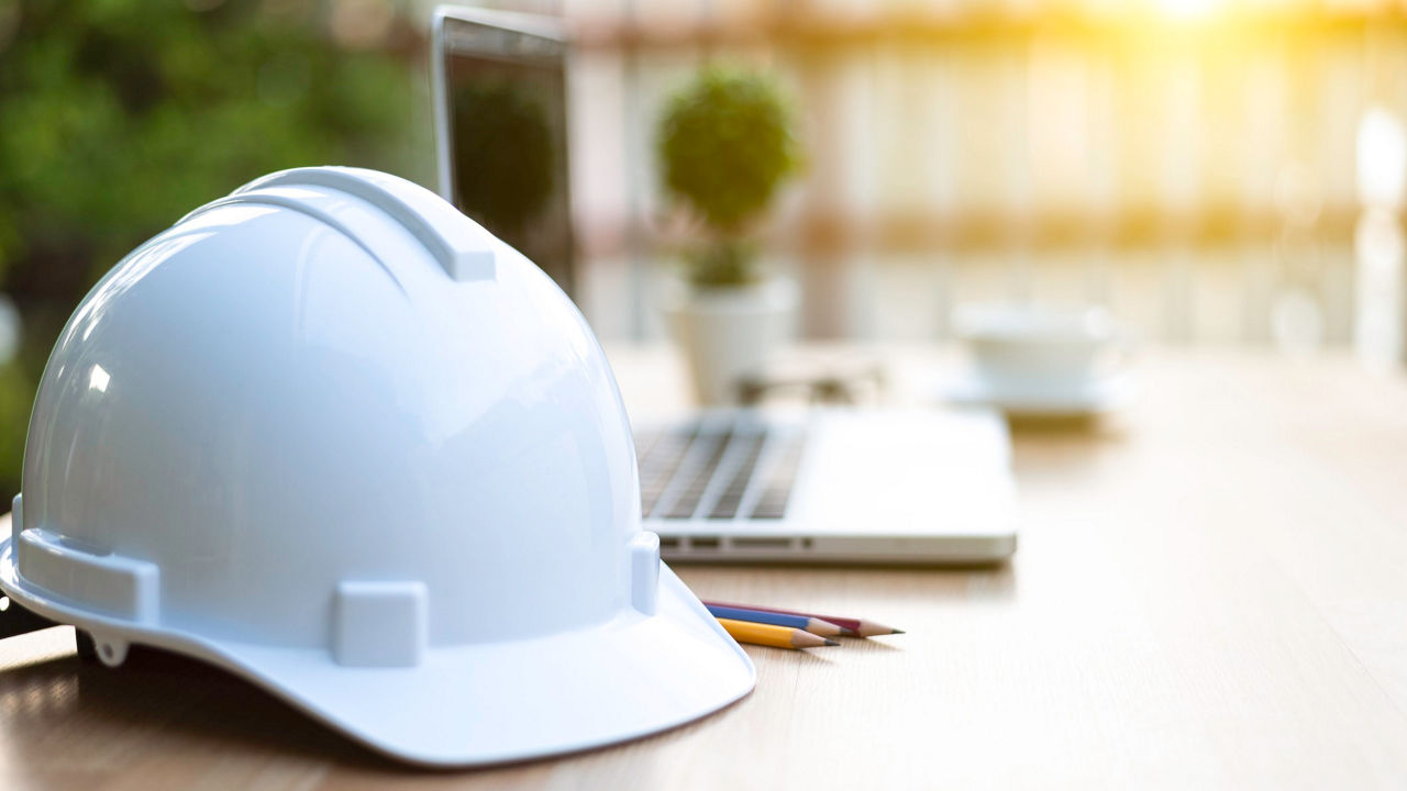 A white hard hat sits on a desk next to a laptop.