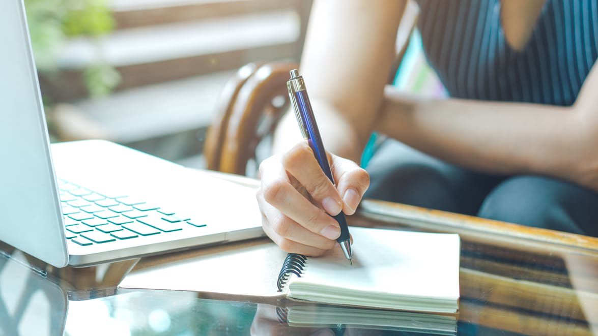 A woman writing on a notebook next to a laptop.