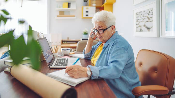 An elderly woman sitting at a desk and talking on the phone.