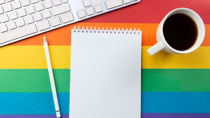 A blank notepad with a cup of coffee and a keyboard on a rainbow colored background.