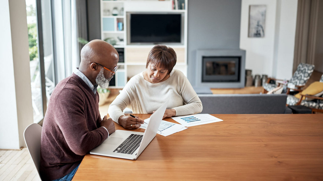 A man and woman working on a laptop at a table in their home.
