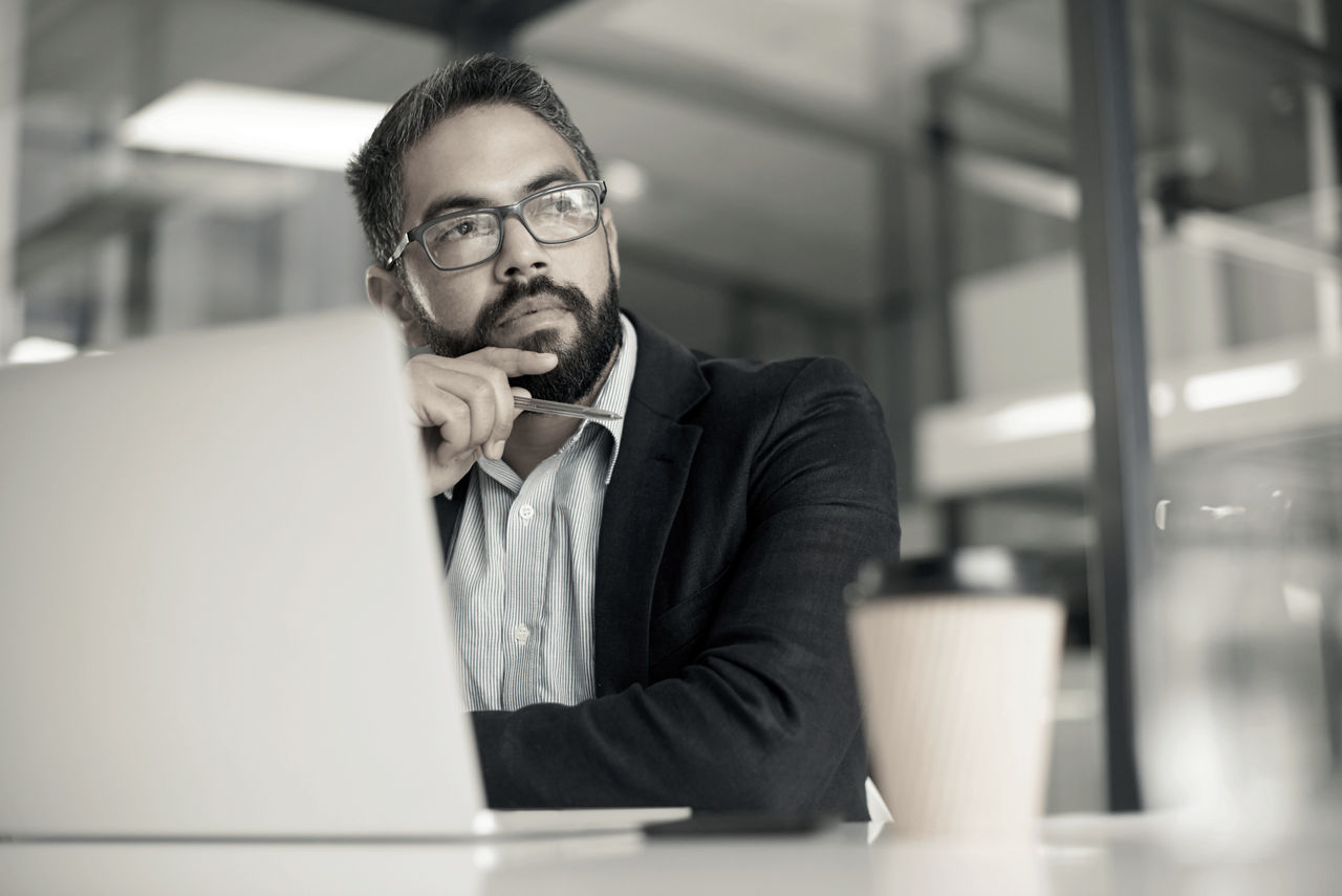 A man sits at a desk with a laptop and coffee cup on it. He is staring off into the distance, as if in thought.