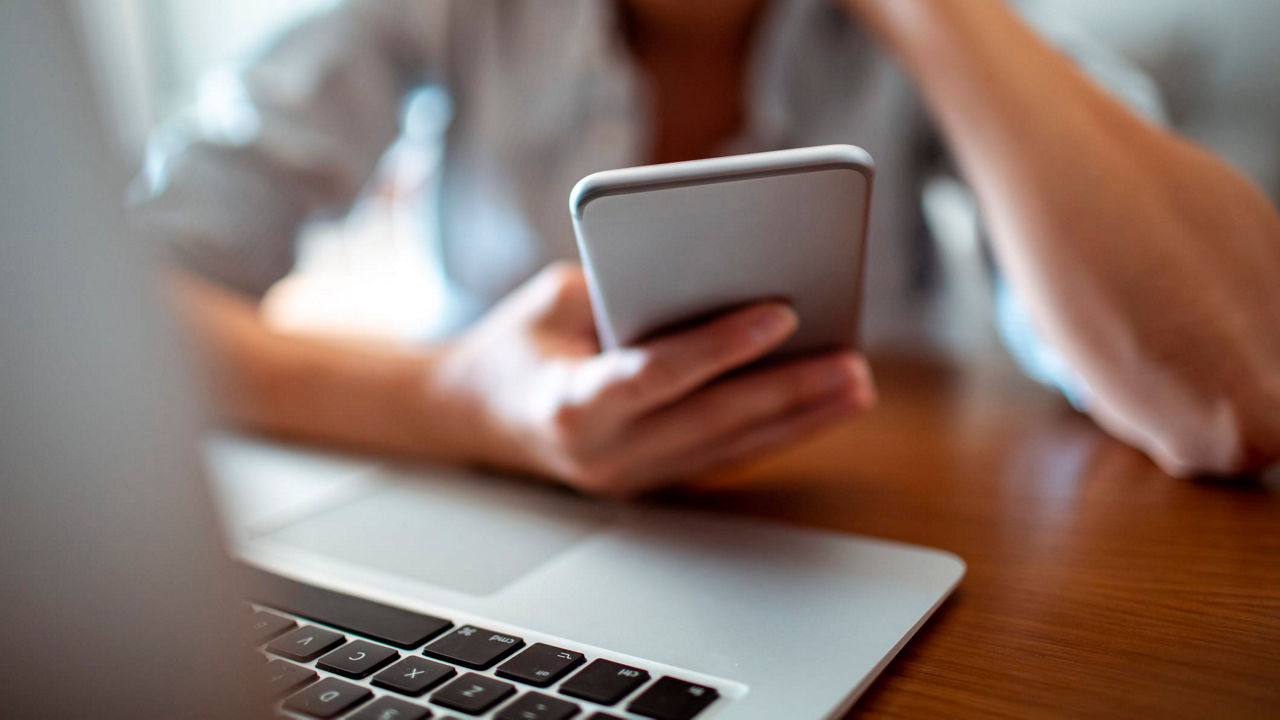A woman is using a cell phone in front of a laptop.