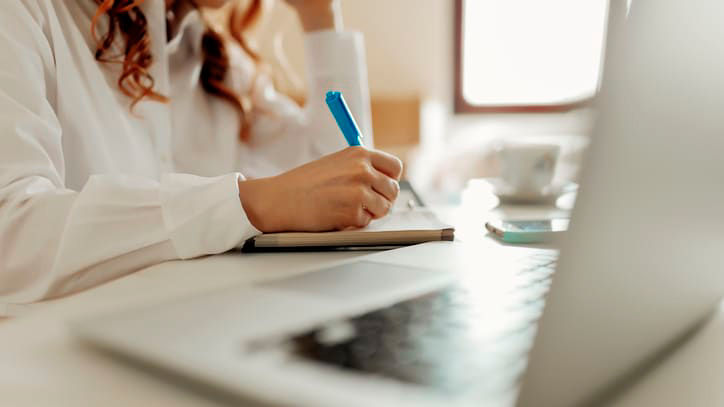 A woman writing in a notebook on a desk with a laptop.